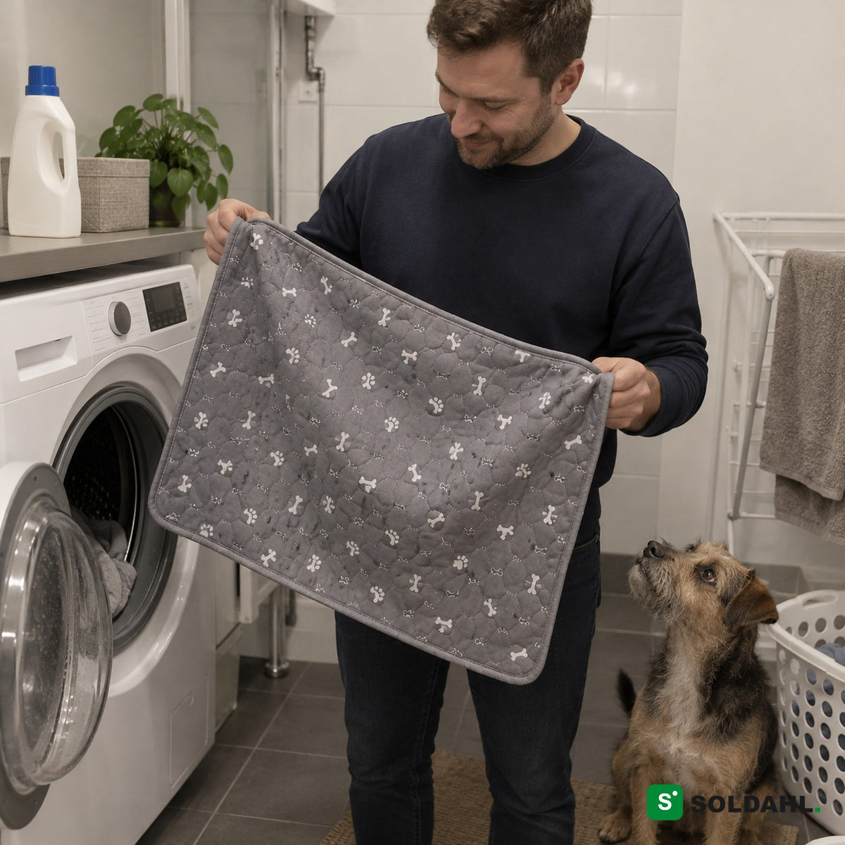 Mand tager den vaskede grå hvalpemåtte ud af vaskemaskinen, mens hunden kigger på